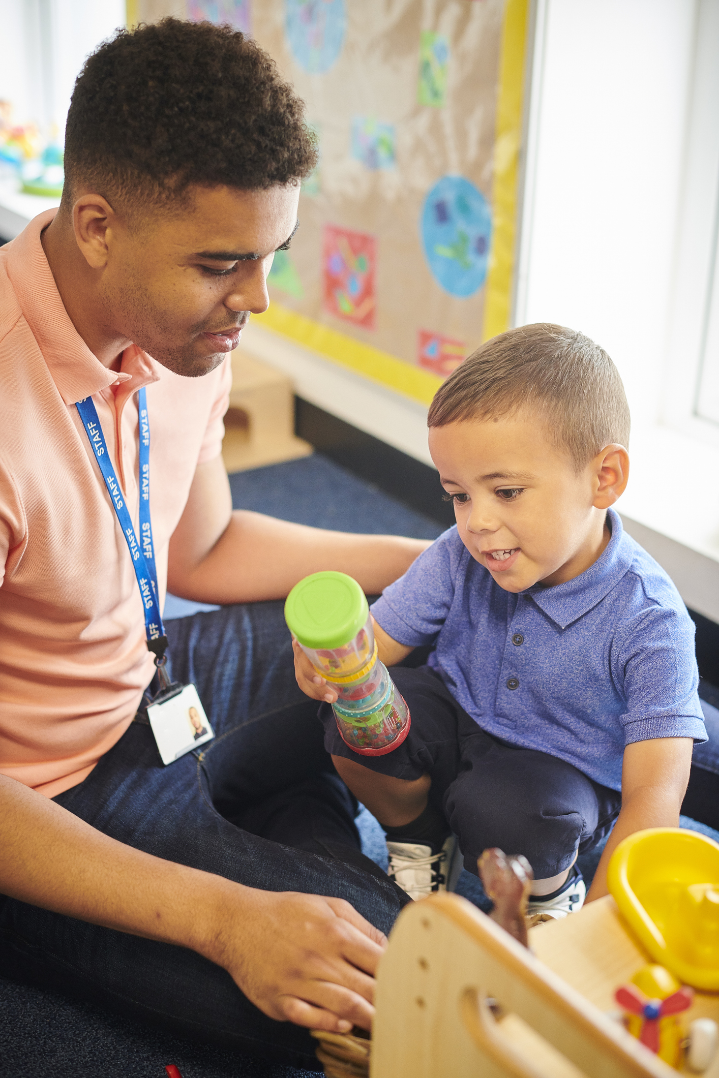An adult sitting on the floor with a young boy holding a green-capped container in a classroom decorated with children’s artwork.