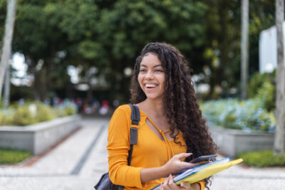 A young woman with long curly hair, wearing an orange top, stands outdoors holding notebooks and a phone, smiling. Trees and a walkway are in the background.