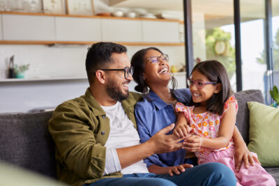 A man, woman, and young girl sit together on a couch in a living room, smiling and laughing.