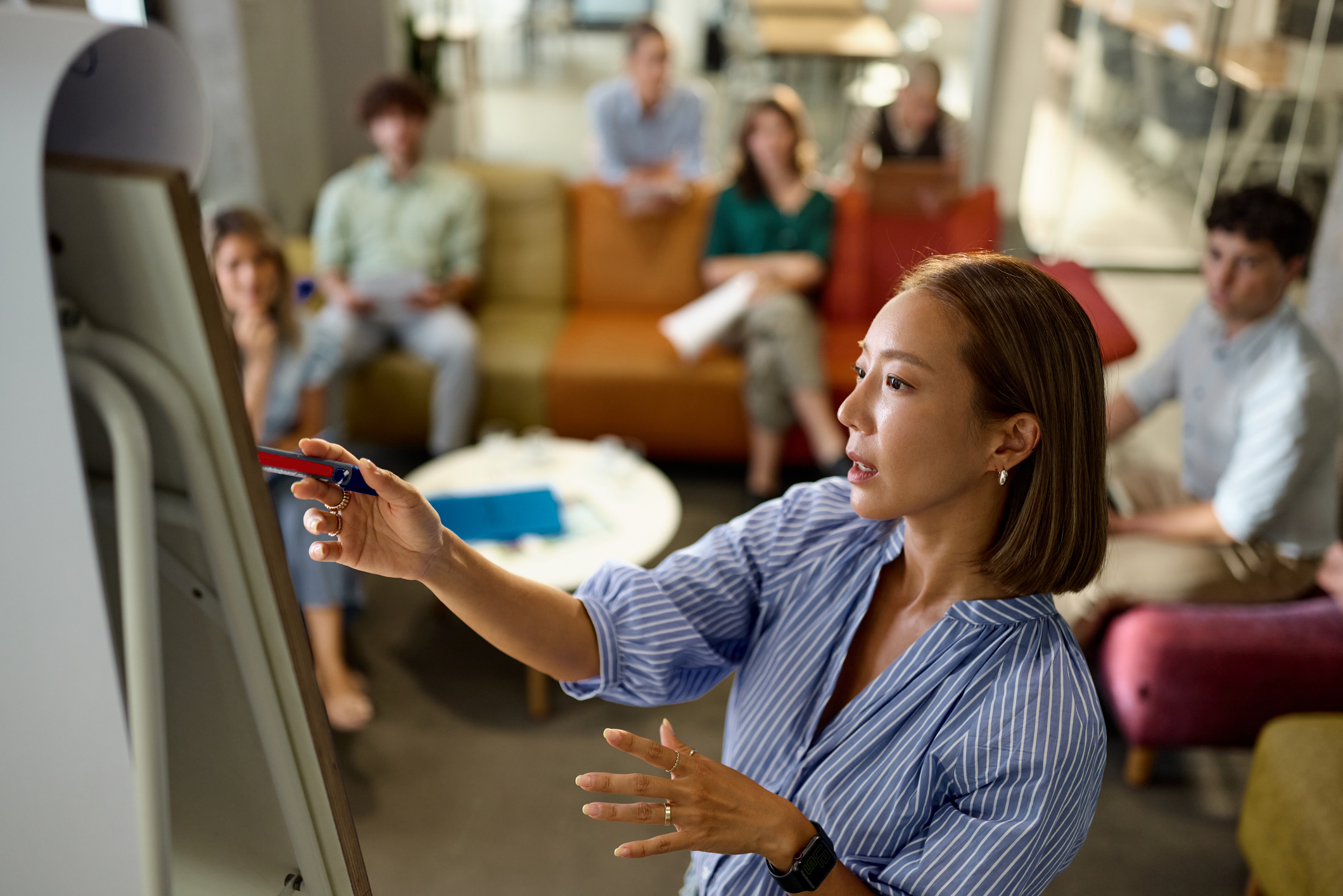 A woman writes on a flipchart while a group of people seated on sofas watch and take notes in a modern office setting.