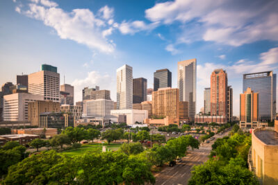 Downtown cityscape with modern high-rise buildings, green spaces, and a partly cloudy sky in the background. Streets and trees are visible in the foreground.