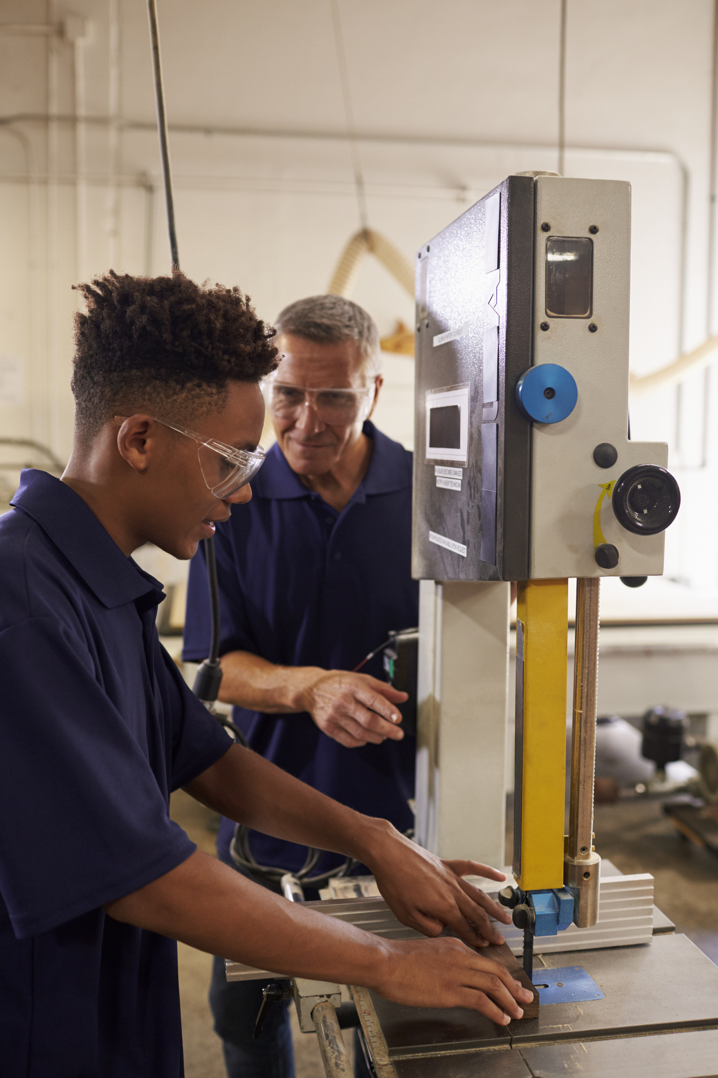 Two men wearing safety glasses operate a bandsaw machine in a workshop. The younger man adjusts the machine while the older man observes and supervises.