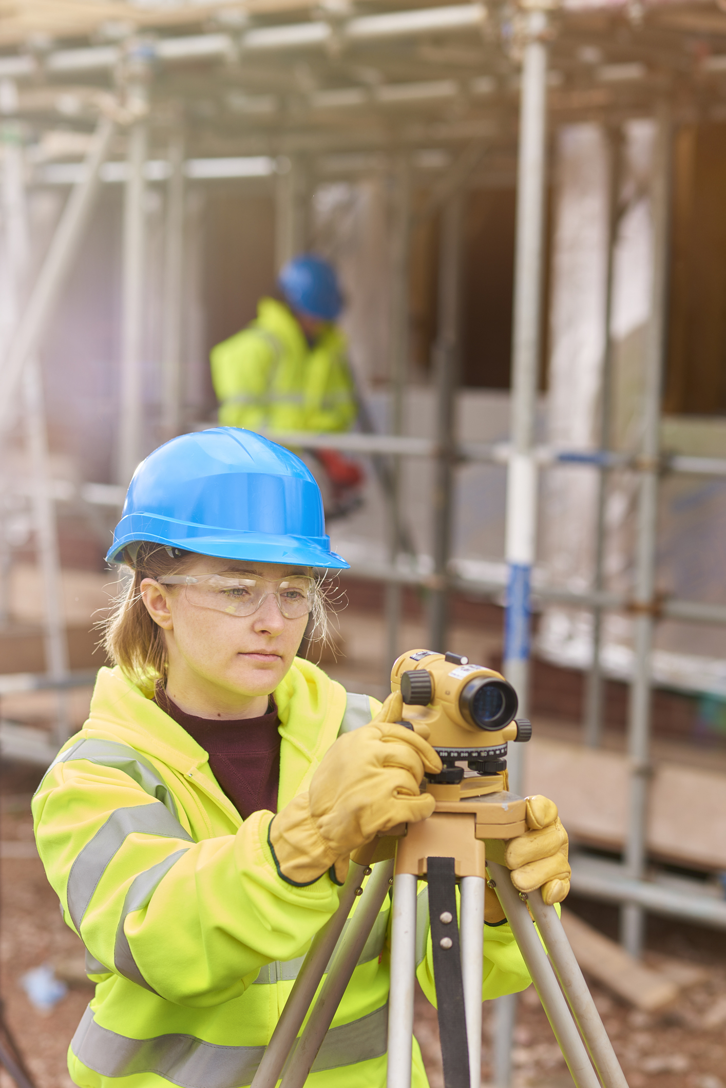 A construction worker wearing a blue hard hat and high-visibility jacket uses a surveying instrument on a tripod at a building site.
