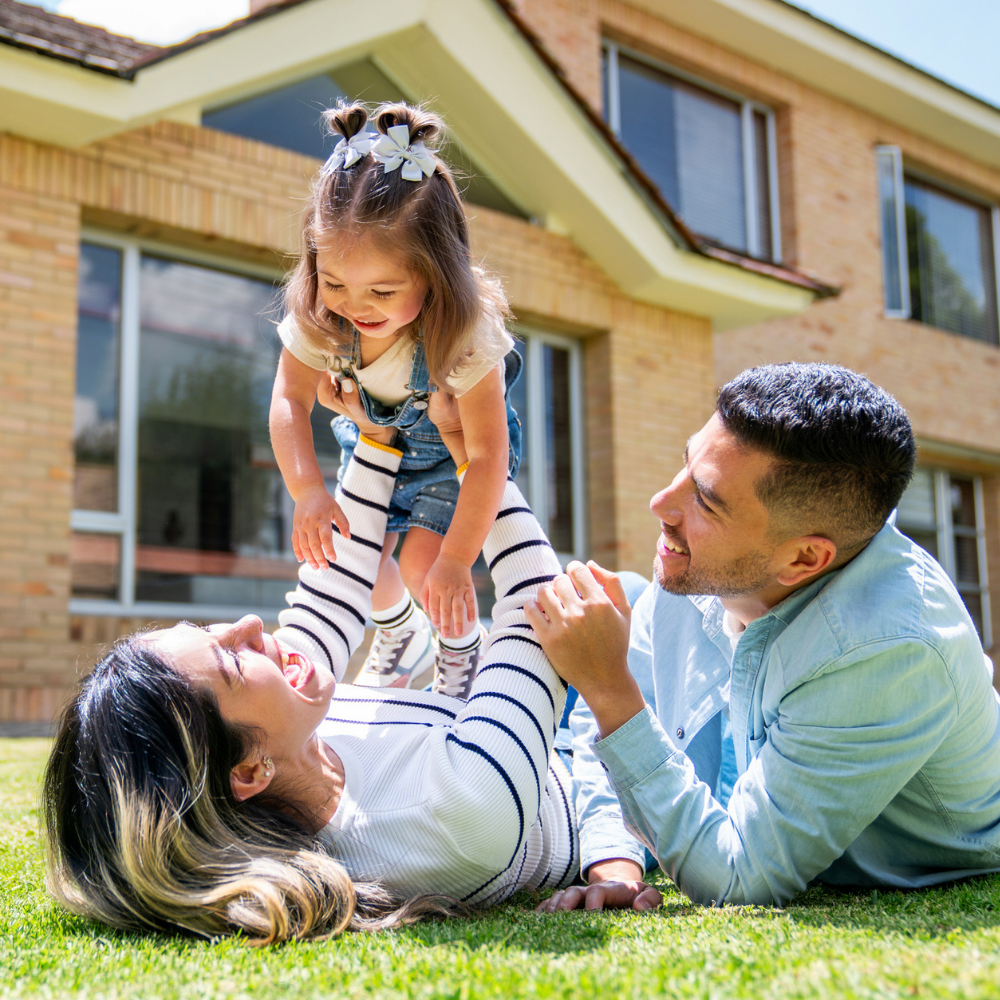 A woman lies on grass holding up a smiling young girl while a man kneels beside them; they are in front of a brick house.