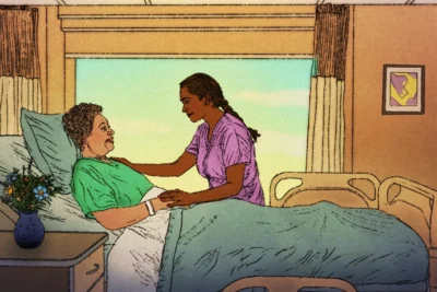 A nurse in purple scrubs stands by a hospital bed, holding hands with a patient who is lying down and smiling. A vase of flowers is on the bedside table.
