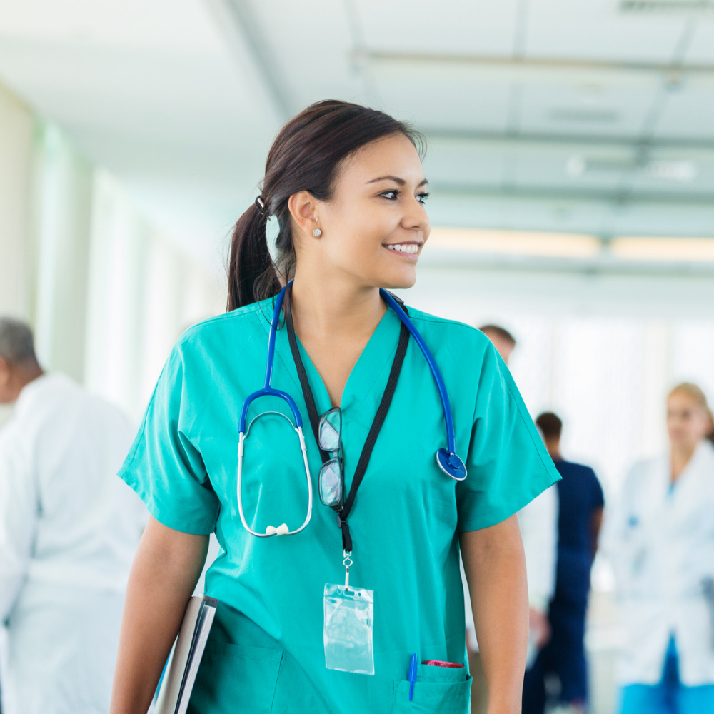 A healthcare worker in green scrubs with a stethoscope around her neck stands in a hospital corridor, smiling, with other staff visible in the background.