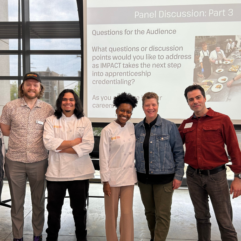 Five people stand together in front of a presentation screen displaying discussion questions at a panel event. Two wear chef coats; the others are in casual attire.