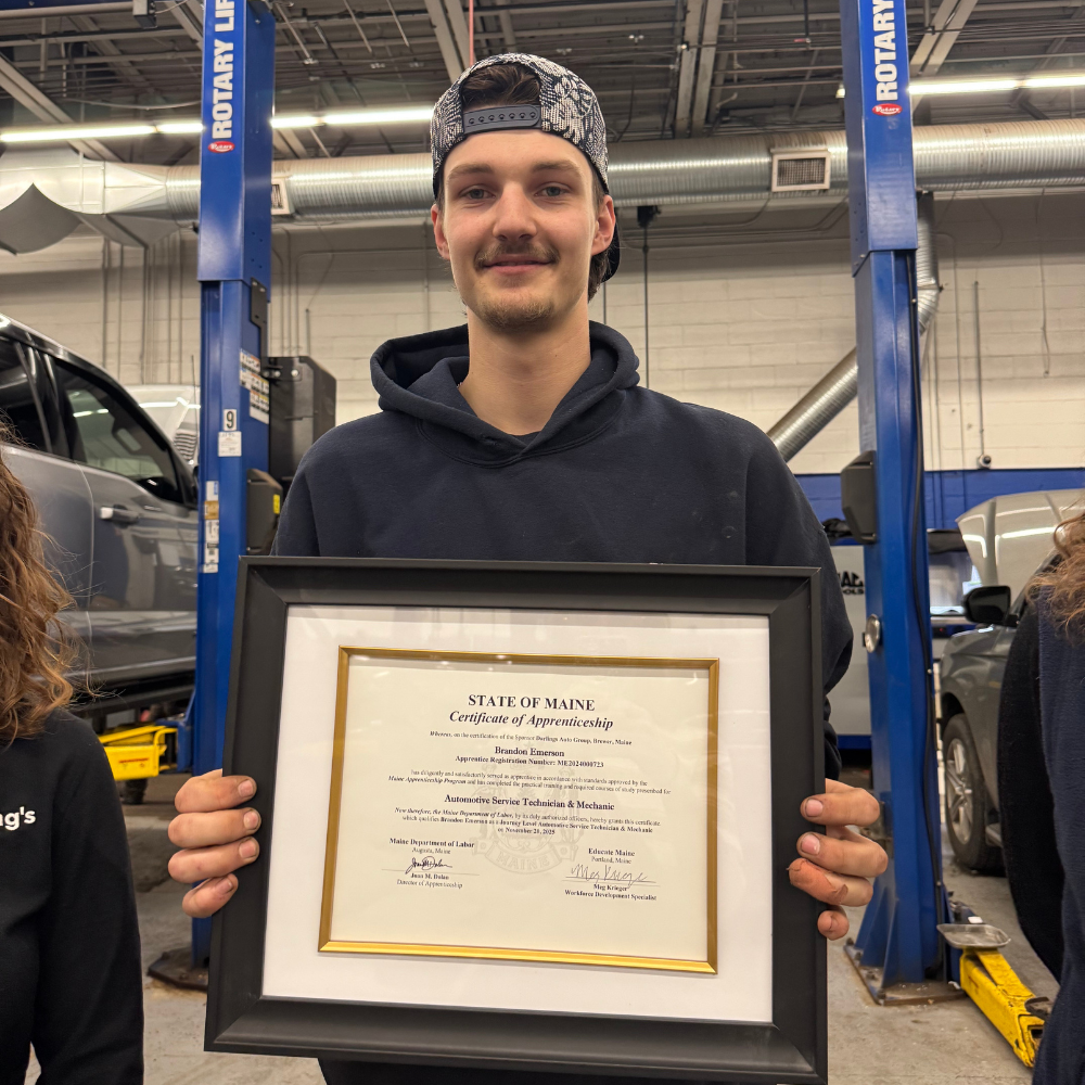 A young man in a workshop holds a framed State of Maine Certificate of Apprenticeship for Automotive Service Technology & Mechanics.
