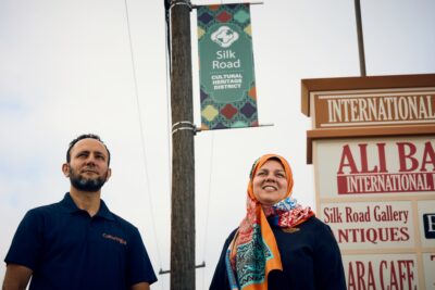 Two people stand near a street sign for the Silk Road Cultural Heritage District and a sign for Ali Baba International Market.