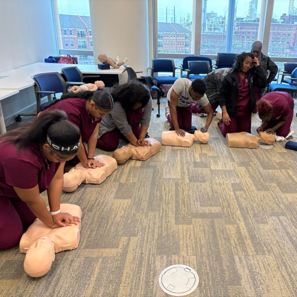 A group of people in scrubs practice chest compressions on CPR manikins in a classroom setting.
