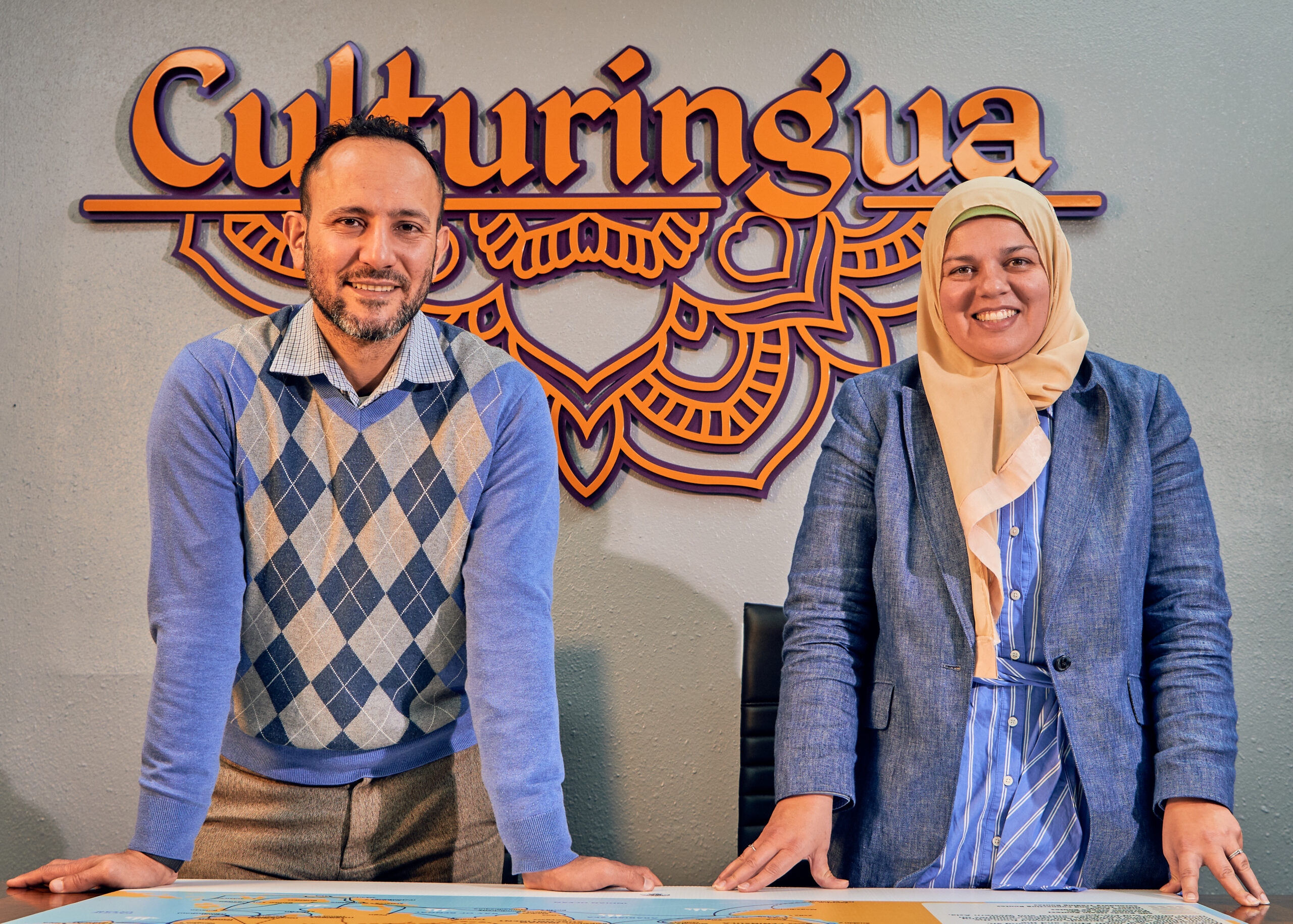 Two people stand side by side at a desk with a map, in front of a wall sign that reads 