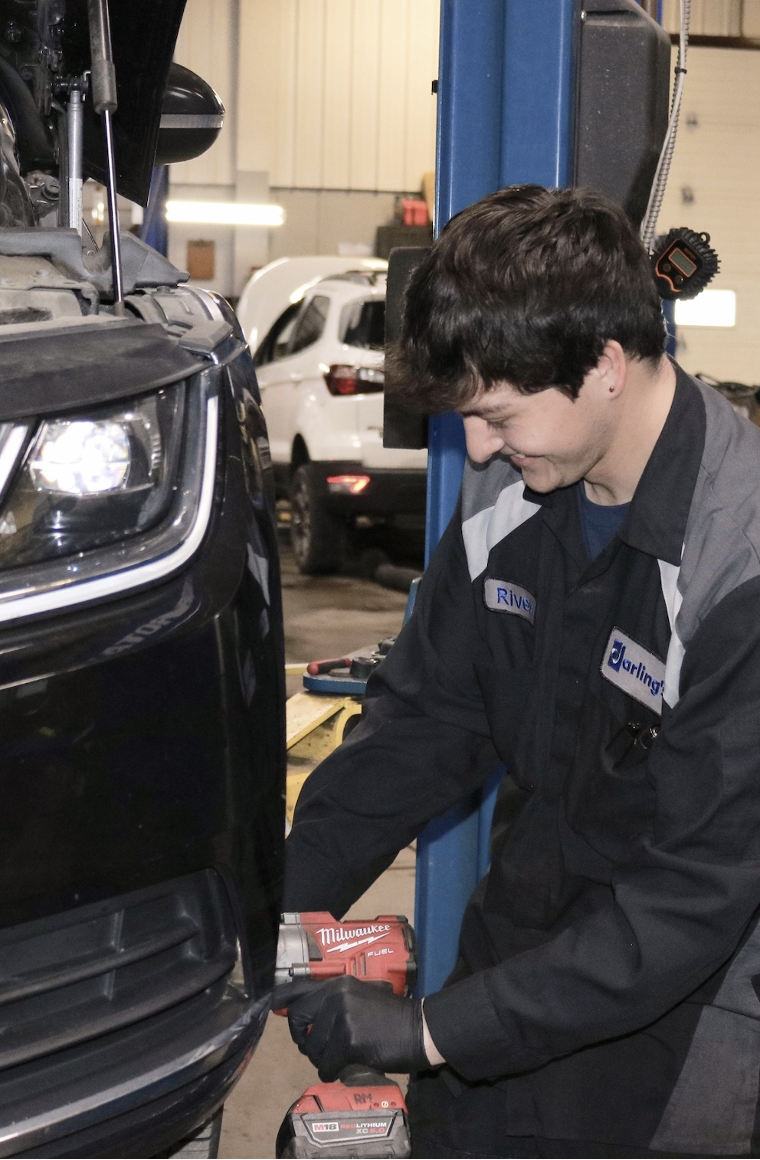 A mechanic in uniform uses a power tool to work on the front of a black vehicle in an auto repair shop.