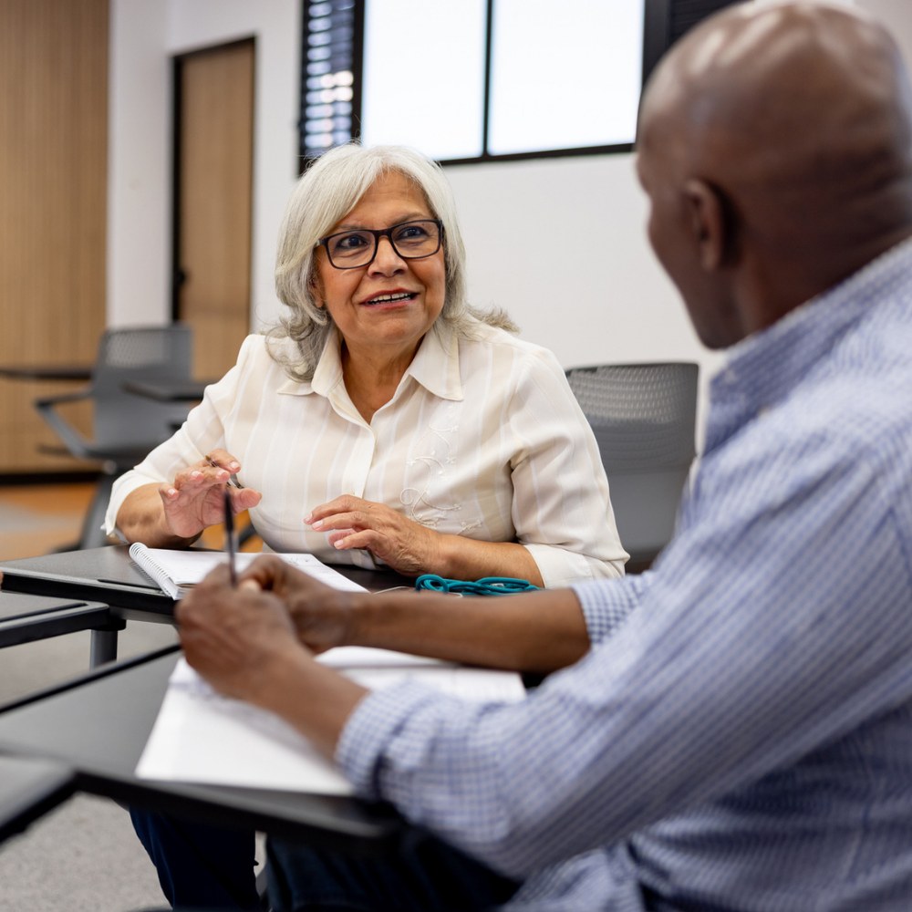 Two older adults sit at desks in a classroom, talking and taking notes on paper with pens.
