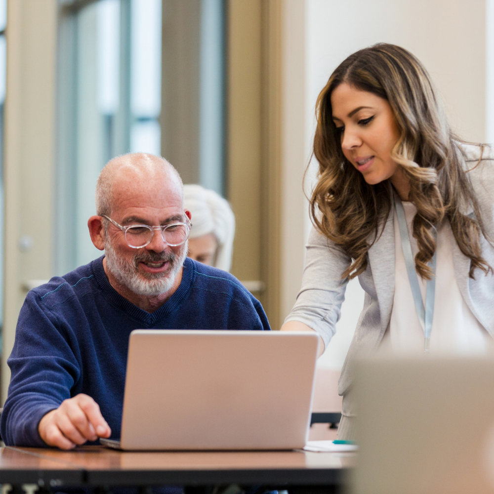 A woman stands beside a seated older man, pointing at his laptop screen while they both look at it in a classroom or office setting.