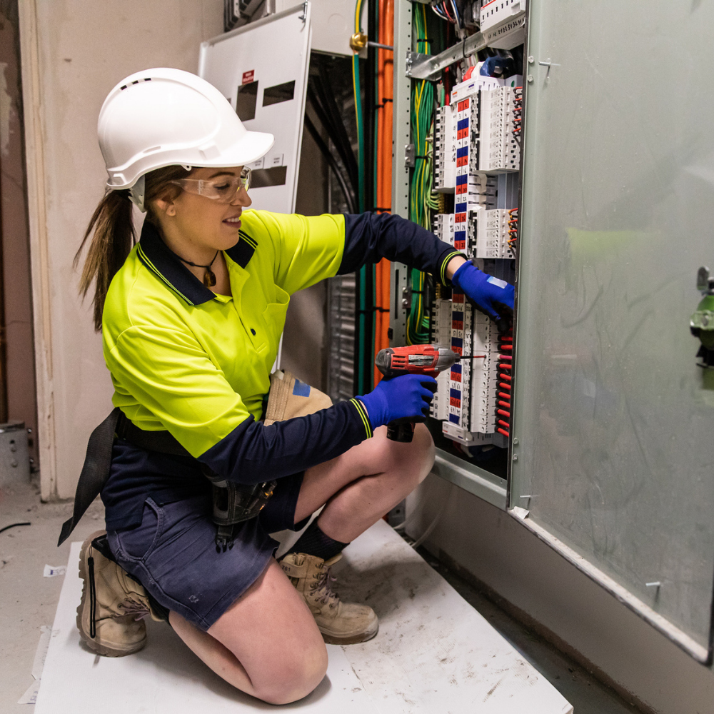 A person wearing safety gear uses a screwdriver to work on an electrical panel with multiple wires and switches in an industrial setting.