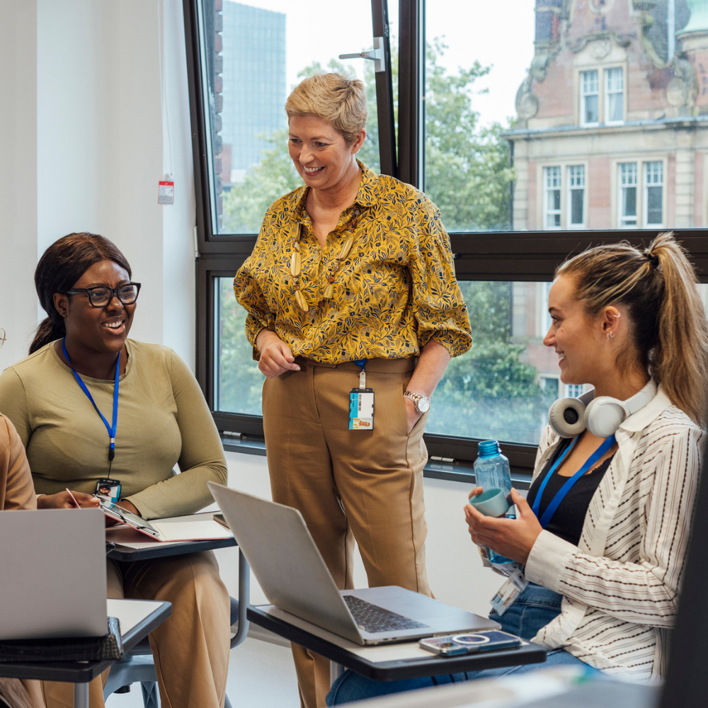 Four women in an office setting are having a discussion, with one woman standing and smiling while the others sit and listen attentively.