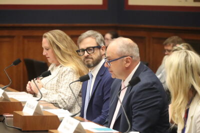 Four people in business attire sit at a panel table with microphones, speaking and listening during a formal meeting or hearing in a wood-paneled room.