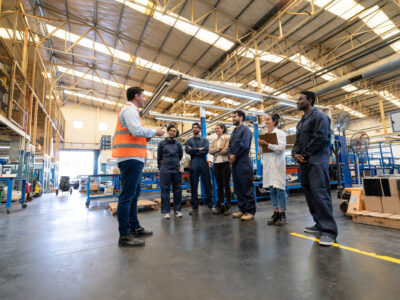 A supervisor in an orange safety vest talks to a group of factory workers standing together on the floor of a large industrial facility.