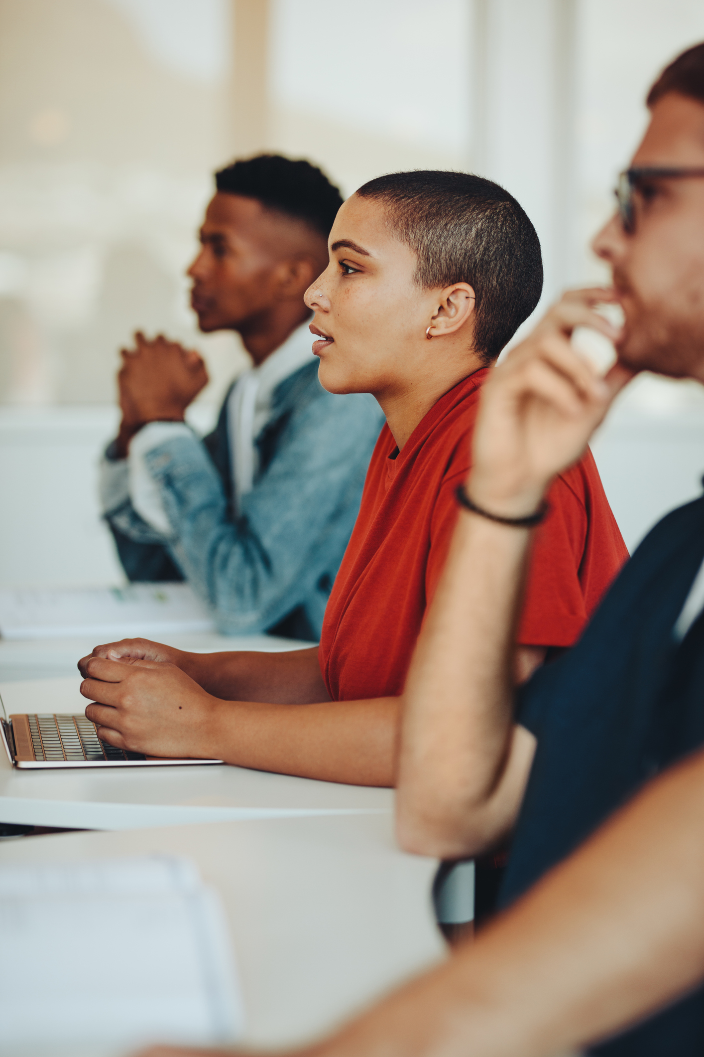 Three students sit at desks in a classroom, focused and attentive, with one using a laptop and others appearing to listen to something out of frame.