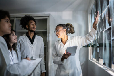 Four scientists in lab coats examine and discuss X-ray images on a light board in a clinical setting.