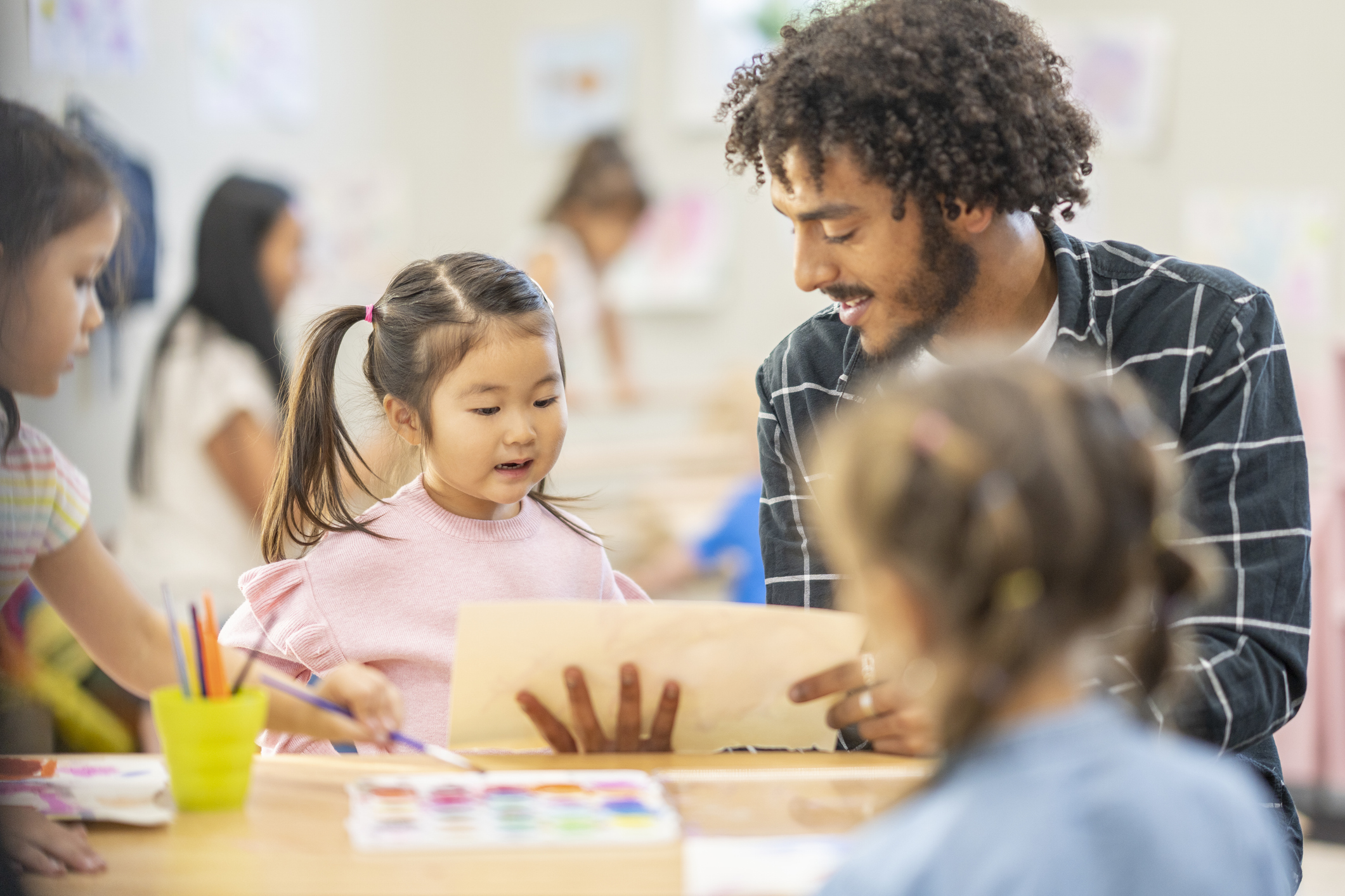 A man and a young girl sit at a table, looking at a piece of paper together, with other children drawing and painting nearby in a classroom.