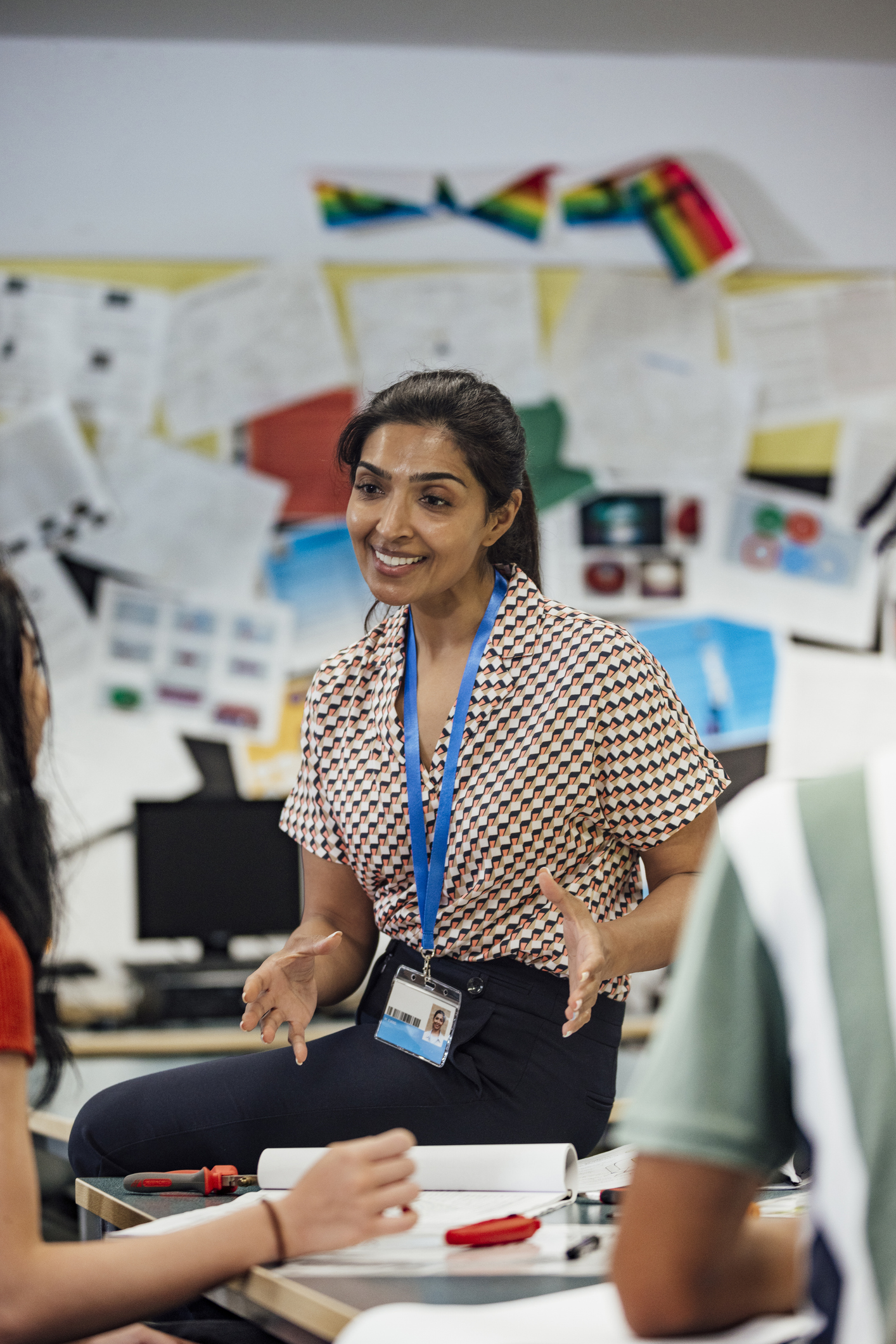 A woman wearing a lanyard speaks to two people at a table in a classroom, with papers and posters displayed on the wall behind her.