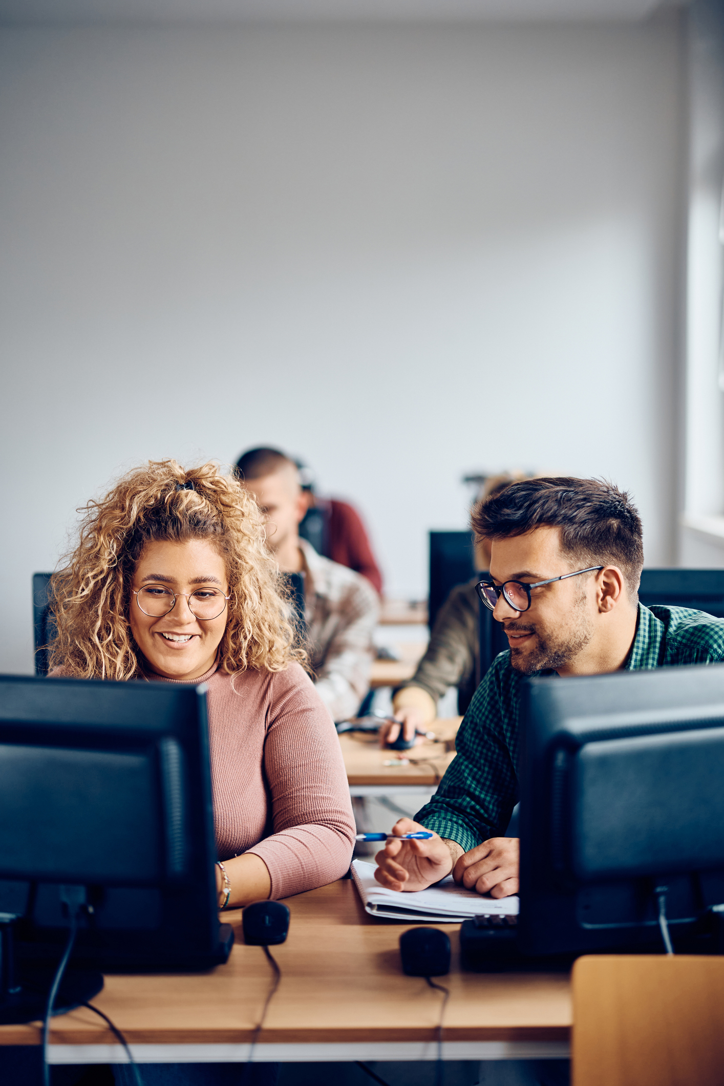 Two people sitting at desks work on desktop computers in a classroom setting, with others working in the background.