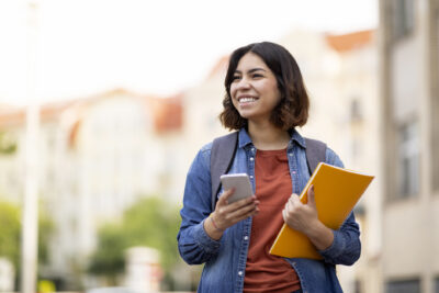 Young woman outdoors holds a smartphone in one hand and yellow notebooks in the other, smiling and wearing a backpack, with buildings blurred in the background.