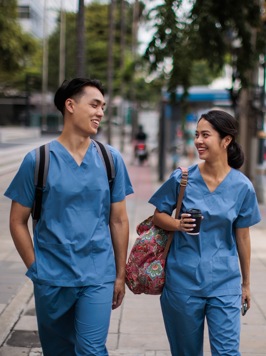 Two people in blue scrubs walk outdoors, carrying bags. One holds a coffee cup. They are smiling and appear to be conversing on a city sidewalk.