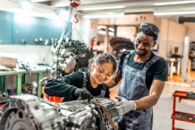 Two mechanics wearing safety goggles and work overalls are working together on a car engine in a well-lit auto repair shop.