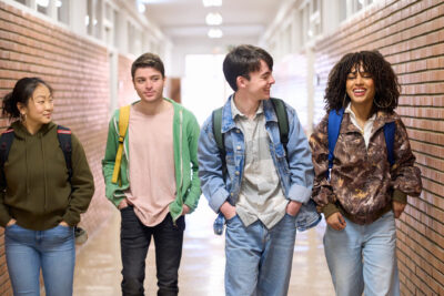 Four students with backpacks walk and talk together in a school hallway lined with brick walls.
