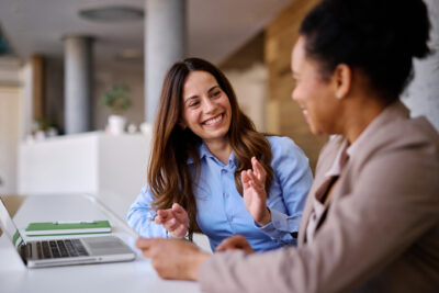 Two smiling businesswomen discussing a project in a modern office, collaborating with a laptop and documents, exemplifying teamwork and effective communication in a professional setting