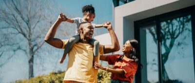 A man carries a young girl on his shoulders while a woman stands beside them, all smiling outdoors near a modern house on a sunny day.