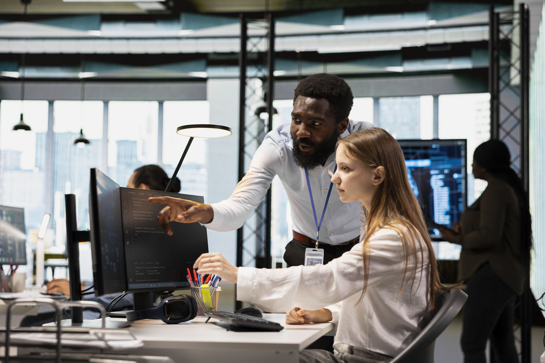 Two colleagues in an office work at a computer; one person is seated and typing while the other stands and points at the monitor, discussing the content shown on the screen.