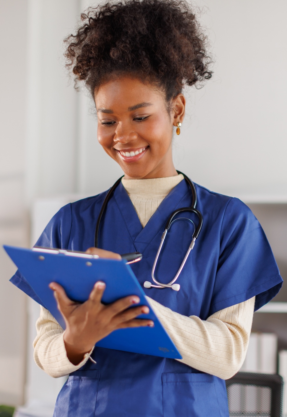A healthcare professional in blue scrubs stands indoors, smiling and writing on a blue clipboard with a stethoscope around their neck.