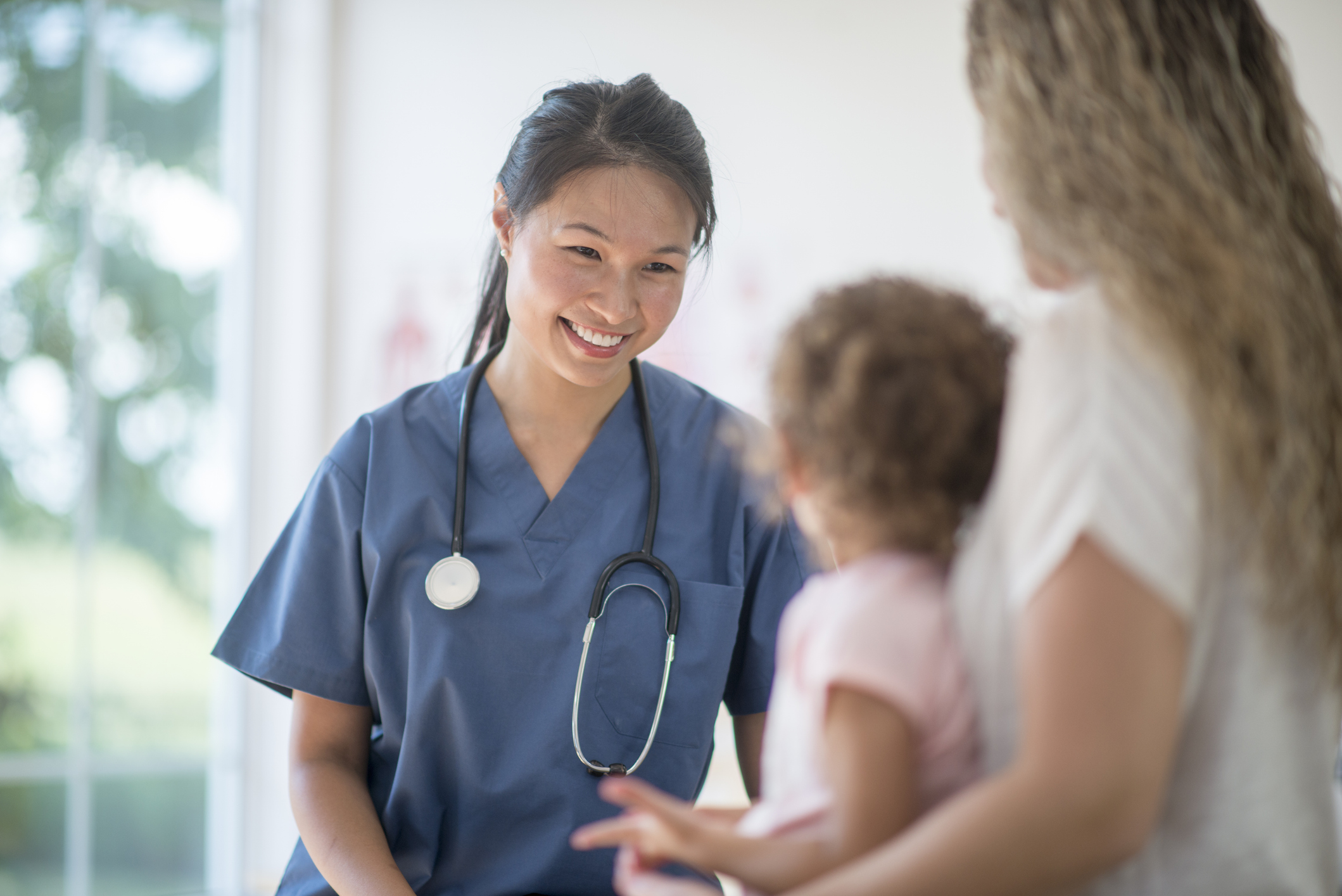 A healthcare worker in scrubs with a stethoscope smiles while talking to a woman holding a young child in a bright medical office.