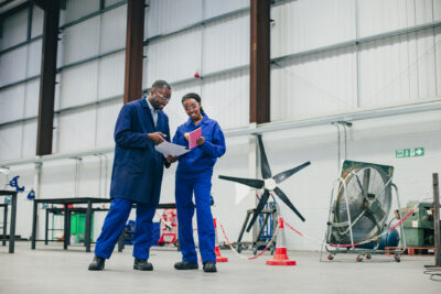 Two engineers in blue coveralls review documents in an industrial workshop, standing near aviation equipment and safety barriers.