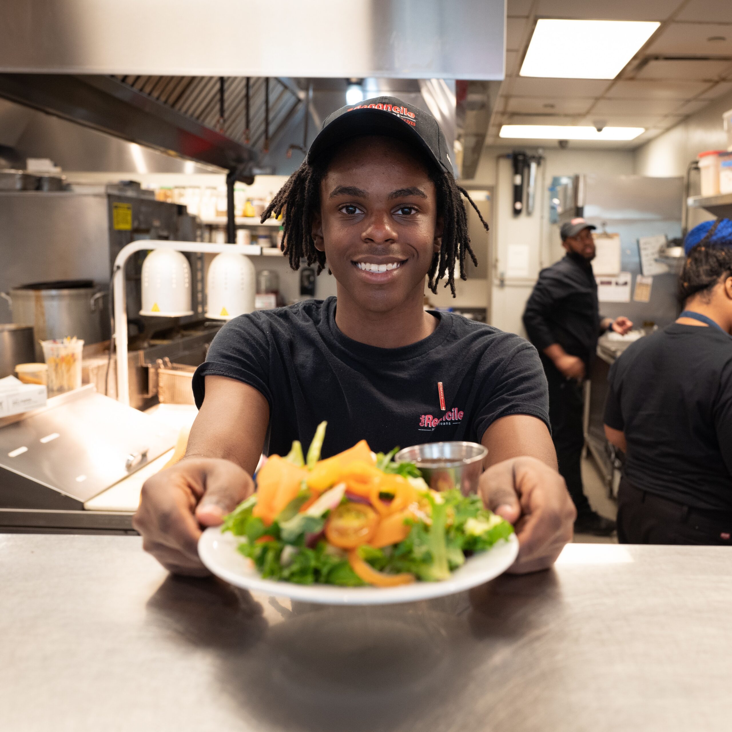 A restaurant worker in uniform smiles and holds out a plate of fresh salad across the counter in a busy kitchen.