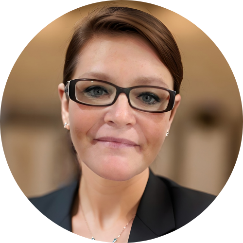 A woman with short brown hair and glasses, wearing a black blazer and a necklace, is facing the camera and smiling slightly.