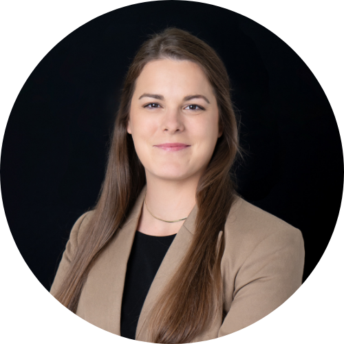 A woman with long brown hair wearing a tan blazer and black shirt poses for a professional headshot against a black background.