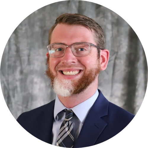 Man with short brown hair, glasses, and a partial white beard, wearing a suit and striped tie, smiling in front of a gray backdrop.