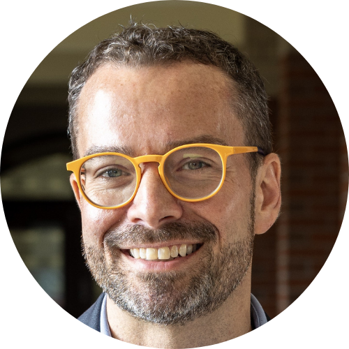 A man with short curly hair, a beard, and yellow glasses is smiling at the camera; background is out of focus.