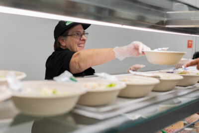 Smiling cafeteria worker hands a bowl of food across the counter, with several filled bowls lined up in front of her.