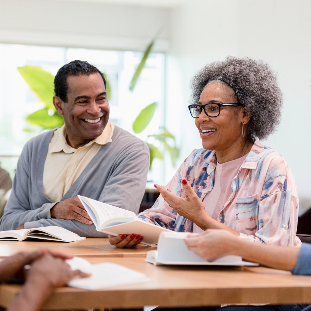Four adults sit at a table, talking and smiling, each holding books, suggesting a book club or group discussion in a bright, casual setting.