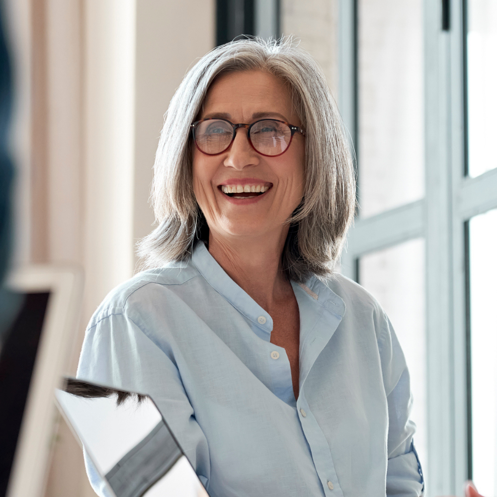 Smiling older woman with gray hair and glasses wearing a light blue shirt, sitting indoors near large windows.