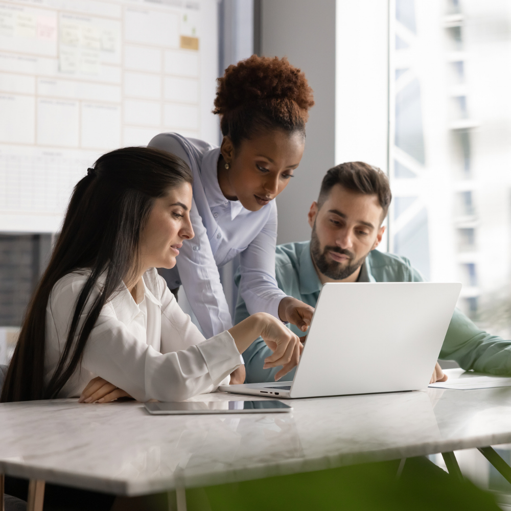 Three people sit at a table in an office, looking at a laptop screen while one person points at it, discussing work together.