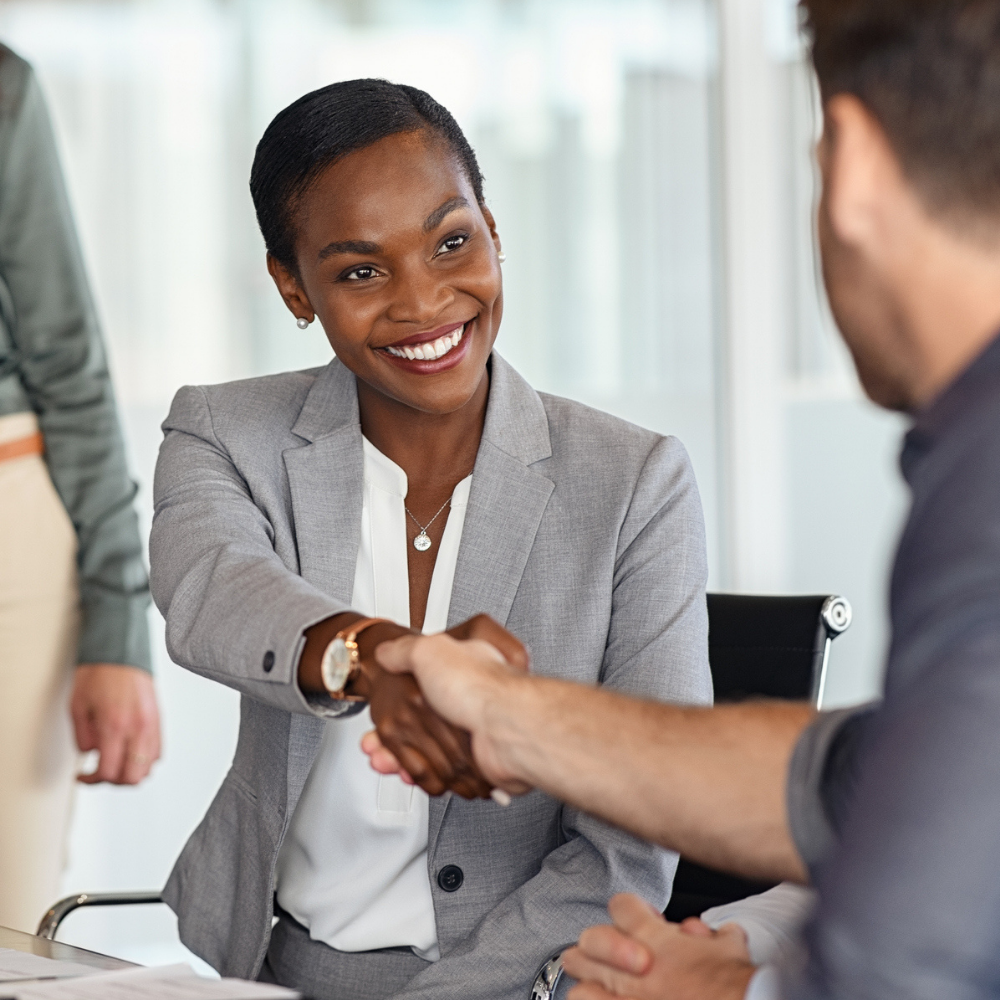 A woman in a gray suit sits at a table, smiling and shaking hands with a man across from her in a professional office setting.