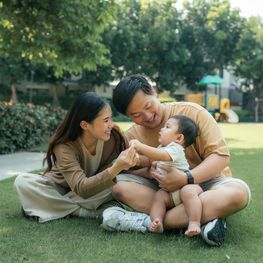A man and woman sit on grass holding a baby. All three are smiling and interacting outdoors, with trees and playground equipment visible in the background.