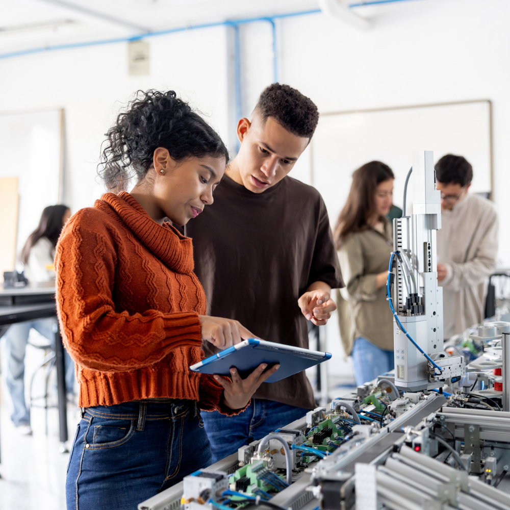 Two students work together in a lab, examining a tablet and electronic equipment. Other people work in the background with various machinery.
