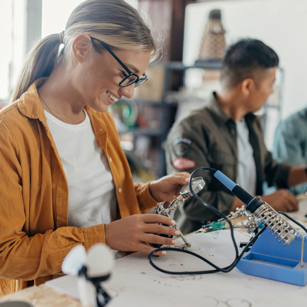 A woman wearing glasses and a yellow shirt uses a soldering iron at a workbench, while another person works in the background.